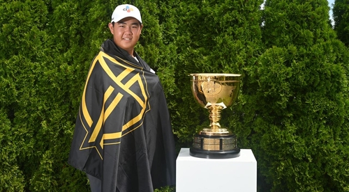 In this photo captured from the official website for the Presidents Cup on Aug. 22, 2022, South Korean golfer Kim Joo-hyung poses next to the winner's trophy while draped in a flag bearing the logo of the International Team. (PHOTO NOT FOR SALE) (Yonhap)